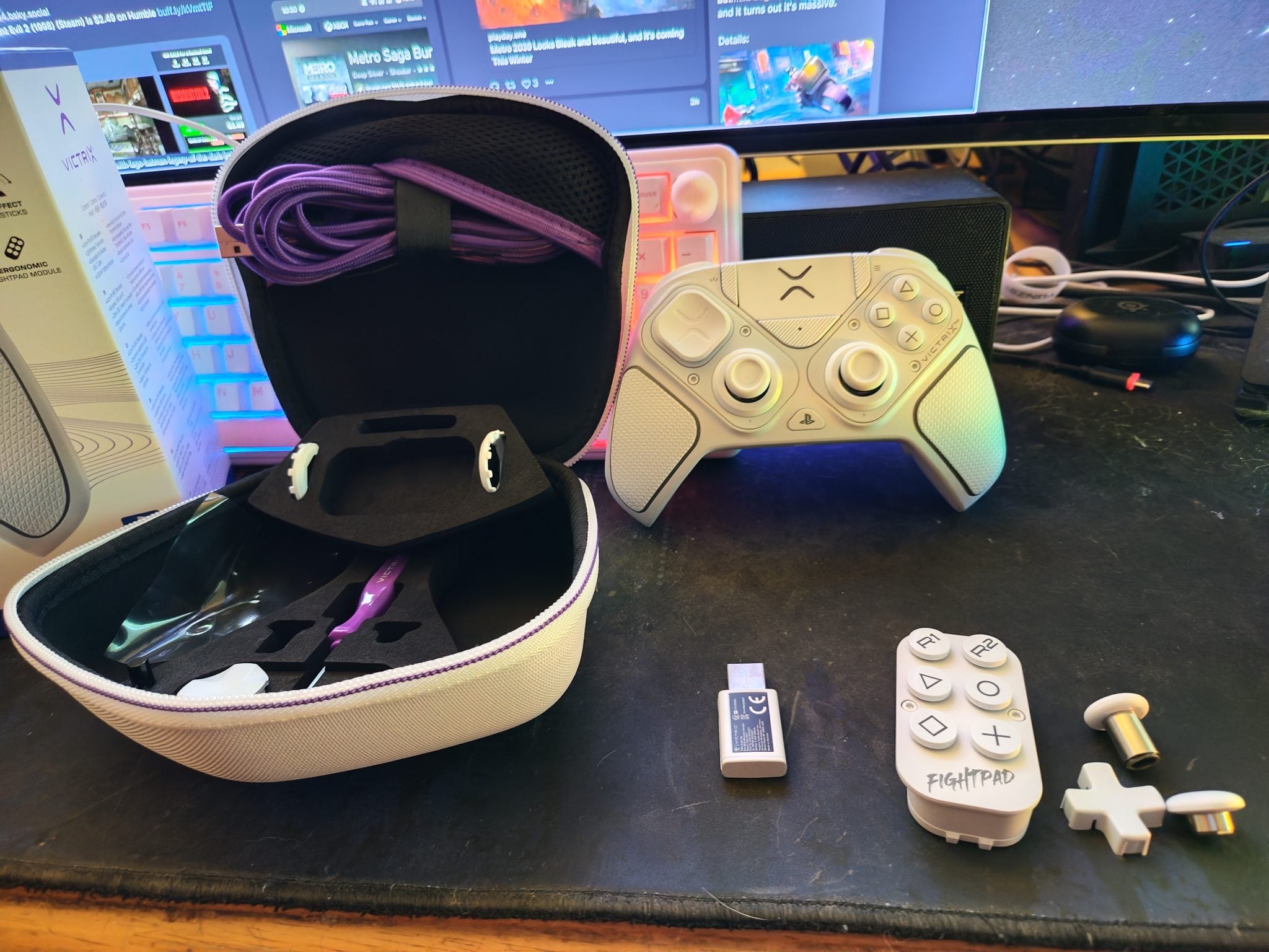 A video game controller in white color sits on a desk next to a black carrying case with a purple cable visible inside. Also shown are additional components like a small wireless adapter and a fight pad with buttons, all on a textured black surface.